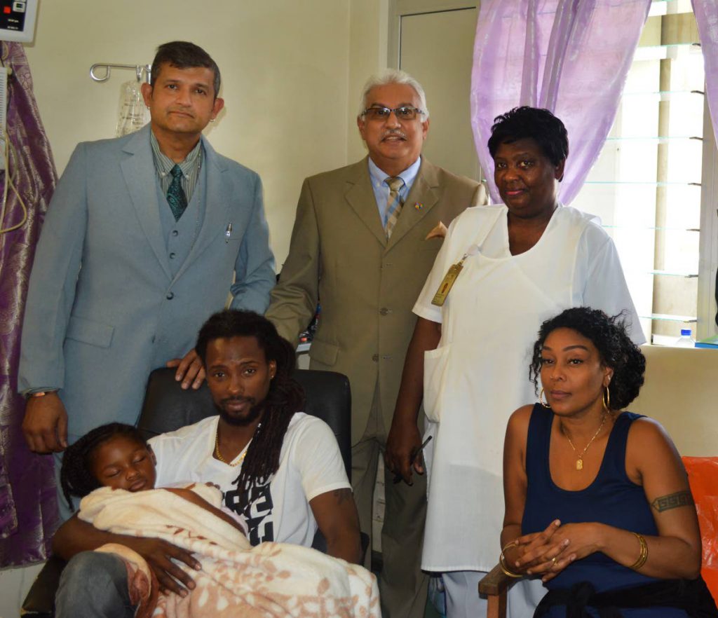 IN GOOD HANDS: Talya Walker, 5, is held by her father David Walker yesterday at the Eric Williams Medical Sciences Complex. Next to them is her mother Trycia Mendoza-Walker.  Behind them, Health Minister Terrence Deyalsingh is flanked by Dr. Gyandeo Maharajh  and a senior nurse.  PHOTO COURTESY MINISTRY OF HEALTH
