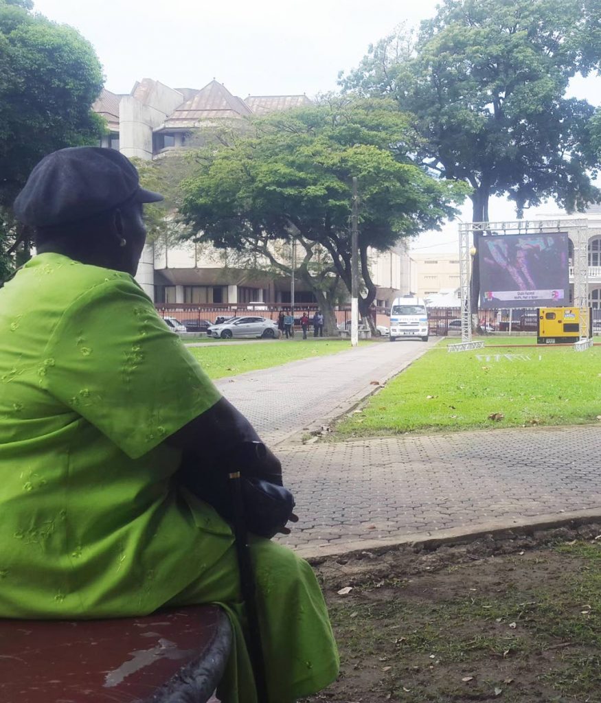 WAITING ON MAX: Phyllis Yallery sits on a stone bench at Woodford Square watching the state funeral of former president George Maxwell Richards, on a screen set up by Government. PHOTO BY KALIFA CLYNE 