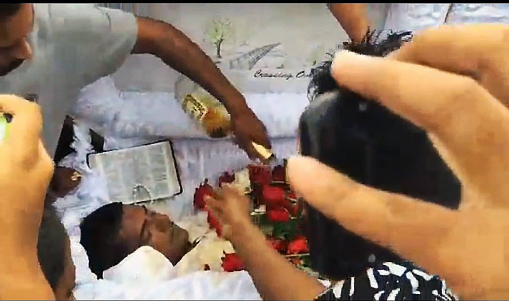 COGNAC SEND-OFF: This screen grab, taken from video images posted to social media, shows a mourner pouring Hennessy cognac on the body of Phillip Basant, brother in law of murdered auto dealer Sheron Sukhdeo. Basant, who himself was gunned down, was cremated on Tuesday.