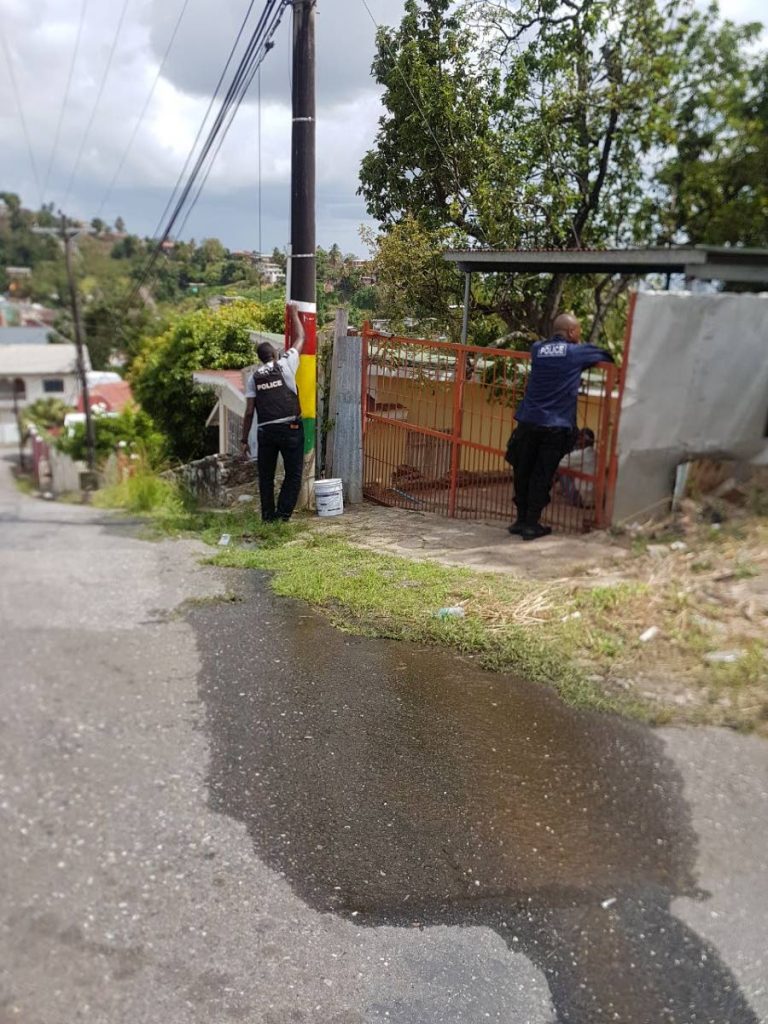 A police  officer attached to the Port of Spain Division re-paints a lamp post defaced by Rasta City gang colours in Gonzales yesterday while another interacts with a resident.