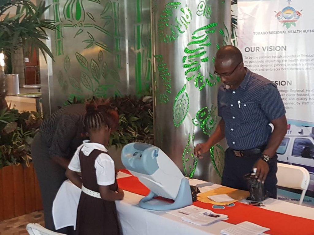 A schoolgirl looks on as Sean Trim of the Blind Welfare Association gets the Visual Acuity Machine ready at the Tobago Regional Health Authority’s (TRHA) ‘Eye Care Everywhere’ Campaign for World Sight Day 2018 at the Gulf City Mall, Lowlands on Monday. 