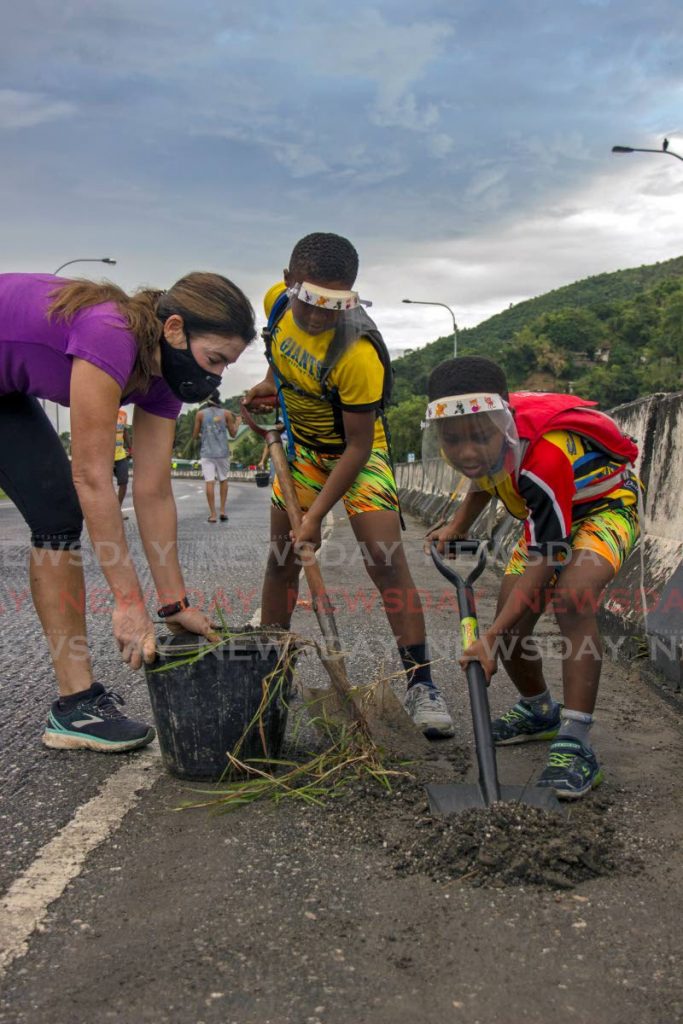 Nathaniel (centre) and Zachai Greaves, nine and six respectively, help Kerry Montano shovel dirt, grass and debris into a bucket as they help to clean a section of the Diego Martin Highway on October 25. A section of the Diego Martin Highway was recently milled to help prevent cars skidding off the road, but which has now made it almost unsuitable for road cyclists. The boys are members of Giants multi-sport club. - Melanie Waithe