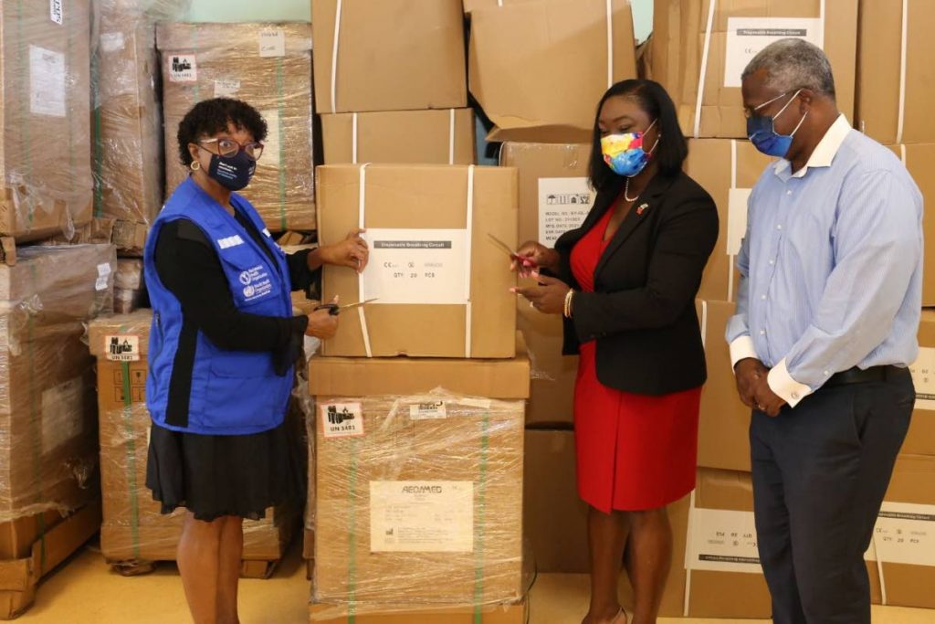 Secretary for Health, Wellness and Family Development, Tracy Davidson-Celestine (centre) snips tie straps along with PAHO country representative Dr Erica Wheeler, left, on one of the boxes filled with ventilator accessories donated by PAHO on Friday. Dr Victor Wheeler, Acting Medical Chief of Staff, TRHA keeps a keen eye on the handover process at the Scarborough hospital.
 - 