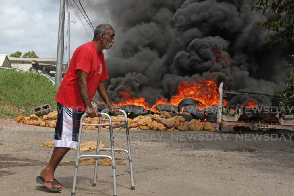 Barrackpore elderly resident, Gadraj Maharaj, 63, carfully crosses Gunness Trace with the aid of a walker during a fiery protests in the community on Monday morning in an attempt to get roads and drainage issues fixed. Photo by Marvin Hamilton
