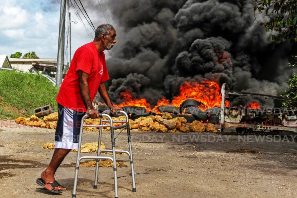 In this July 25, file photo, Gadraj Maharaj, 63, walks near burning tyres and boulders strewn across Gunness Trace, Barrackpore where esidents staged a protest over dilapidated road conditions. Photo by Marvin Hamilton