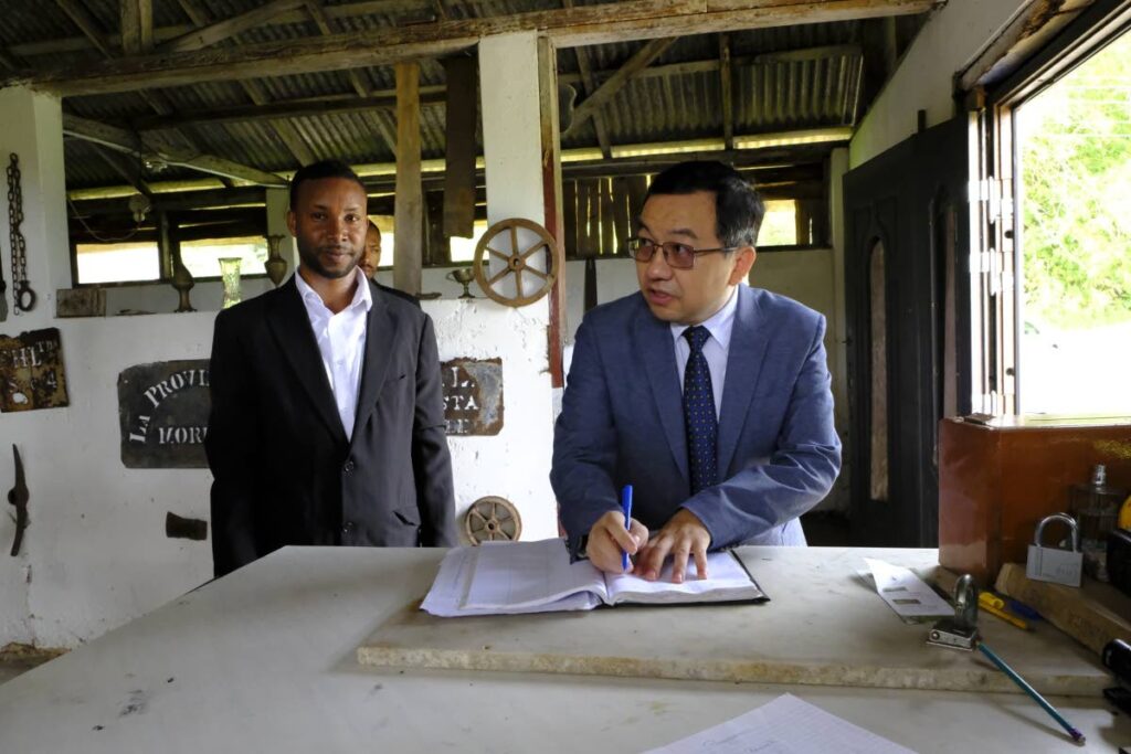 Eric Lewis, left, looks on as Chinese Ambassador Fang Qiu signs the guest book at the Cocoa and Chocolate Museum in Moruga. - 