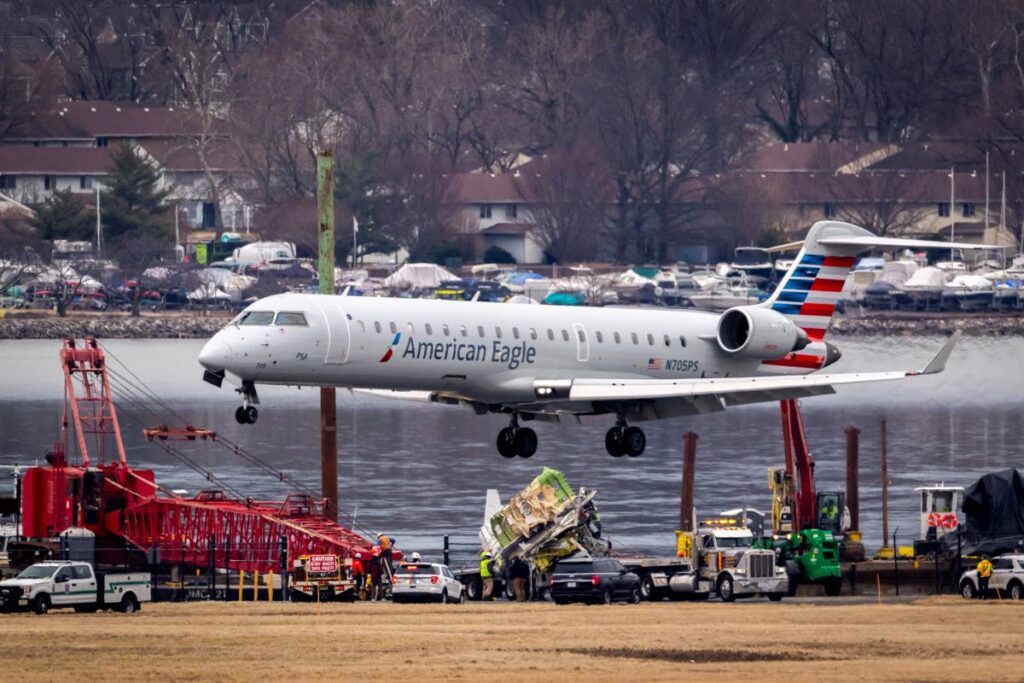 An aircraft lands above a salvaged piece of wreckage on a flatbed truck, near the wreckage site in the Potomac River of a mid-air collision between an American Airlines jet and a Black Hawk helicopter, at Ronald Reagan Washington National Airport, on February 5, in Arlington, Virginia. AP Photo - 